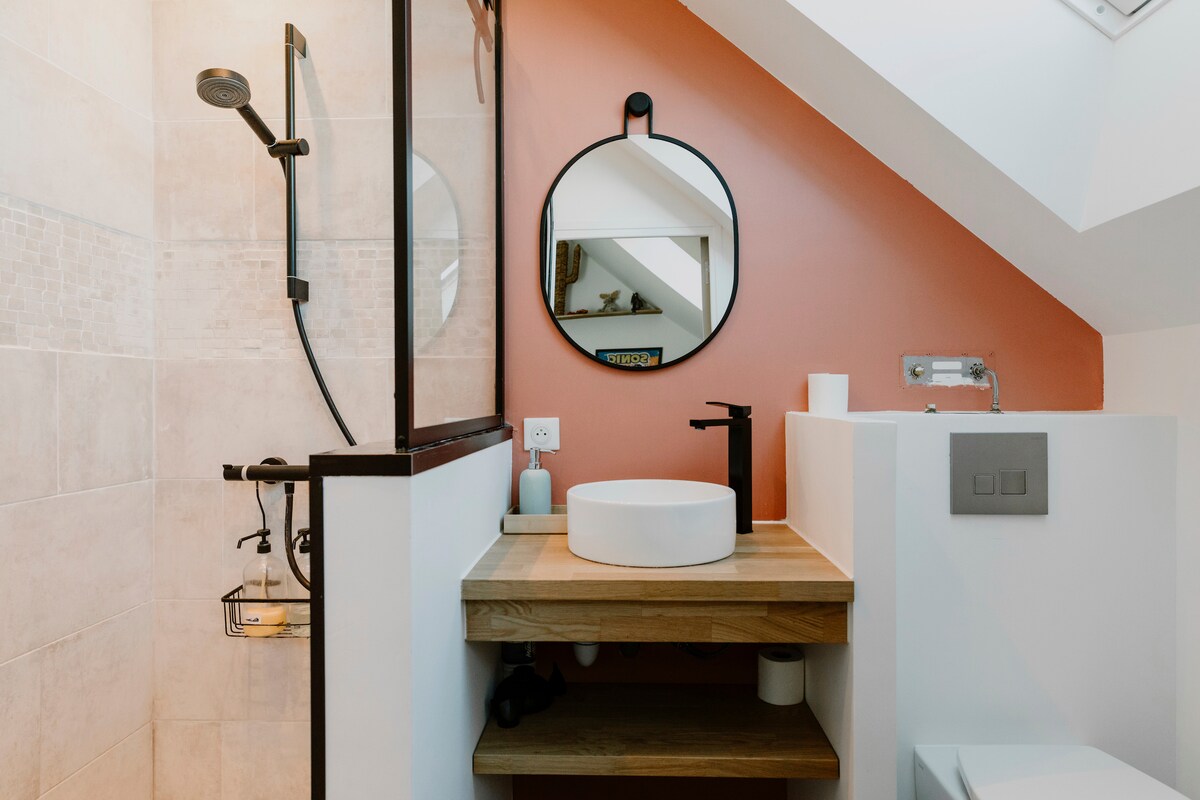 The bathroom features a modern design with light-colored tiles and a contrasting accent wall. A round mirror is mounted above a wooden sink countertop, which holds a circular basin. A walk-in shower with a sleek showerhead and a glass partition adds a contemporary touch.