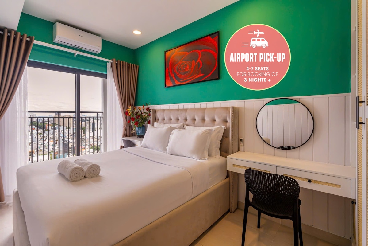 A cozy bedroom is adorned with a vibrant green wall and a striking piece of artwork. A queen-size bed is neatly presented with crisp white linens and rolled towels. Natural light enters through the large window, which offers a view of the city skyline.