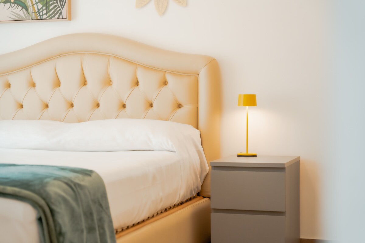 A spacious bed with a tufted headboard is positioned against a light wall. A neatly made duvet and a soft throw are placed at the foot of the bed. A small yellow lamp sits on a modern gray nightstand, providing a touch of color.