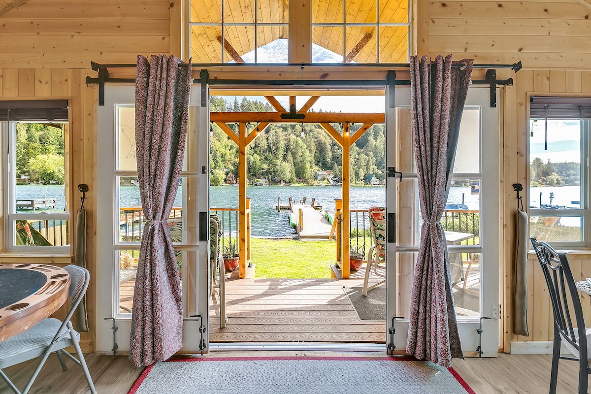 Large double doors open to a sunlit deck overlooking a serene lake, framed by scenic greenery. The inviting outdoor space features a pergola, outdoor seating, and a dock, enhancing the connection between indoor and outdoor living.