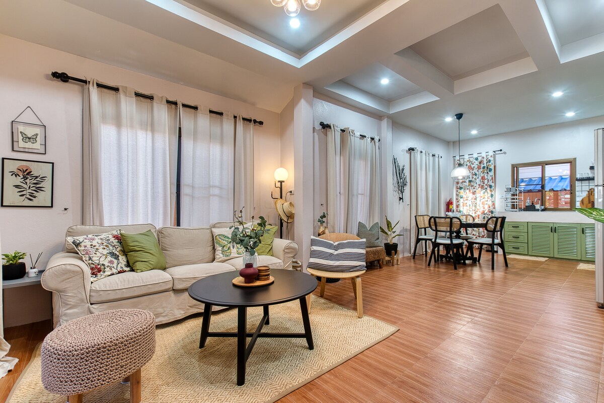 A spacious living area features a comfortable sofa adorned with decorative pillows and a round coffee table at its center. Natural light floods through large windows covered by light curtains. A dining table is visible in the background, accompanied by stylish chairs and a well-organized kitchen.