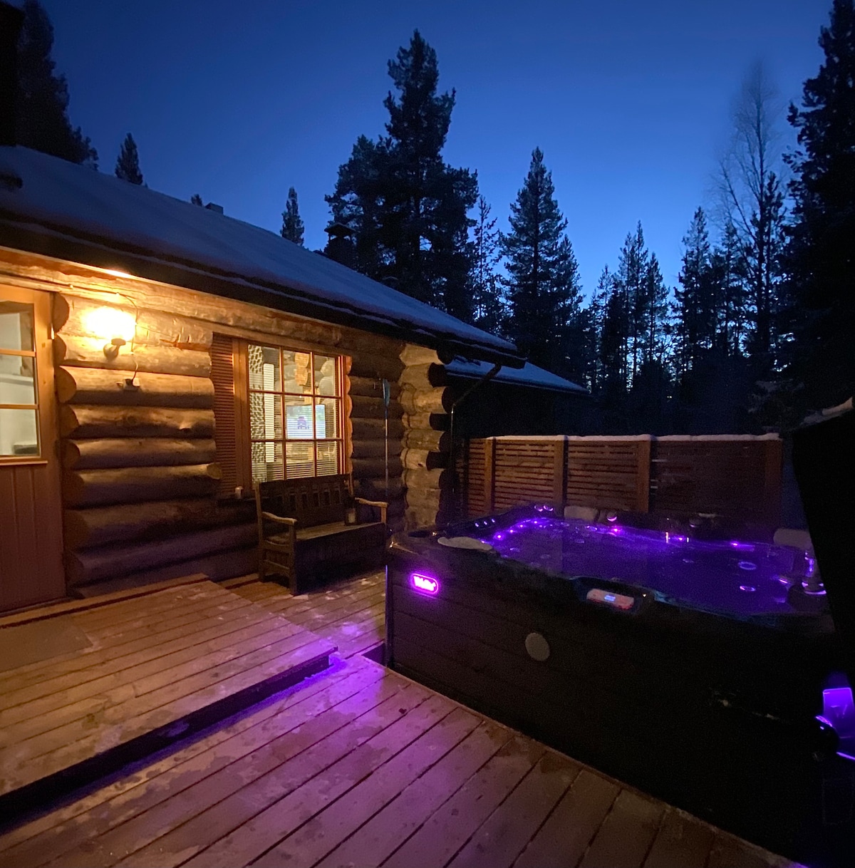 An outdoor terrace is illuminated by soft purple lights, featuring a hot tub beside a wooden structure. Trees surround the area, and a warm glow emanates from a window, enhancing the serene ambiance of the setting.
