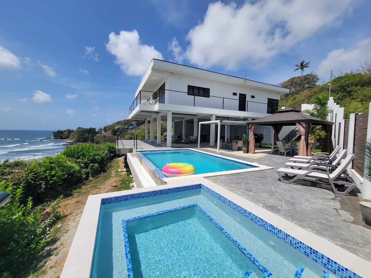 An outdoor area showcases a luxurious saltwater pool accompanied by a hot tub, both with stunning ocean views. A colorful float rests in the pool while sun loungers are arranged nearby. The modern villa structure boasts a sleek design under a bright sky with scattered clouds.