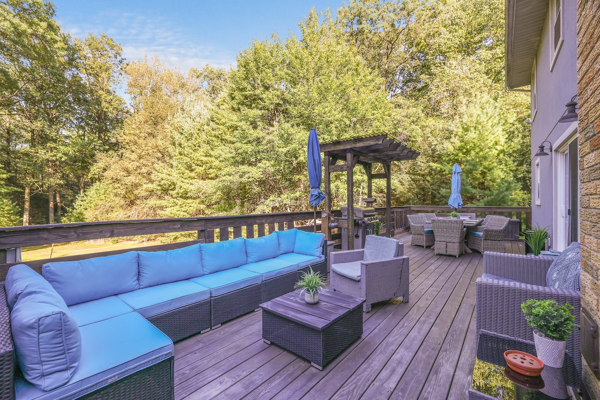 A spacious outdoor deck is shown, featuring weather-resistant lounge furniture in a calming blue tone. There are several seating areas arranged around a central table, with lush greenery and trees visible in the background, creating a serene outdoor environment.