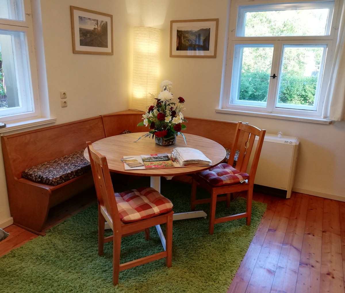 A dining area features a round wooden table surrounded by two wooden chairs with checkered cushions. A cozy corner bench is equipped with a patterned seat and a decorative floral arrangement sits at the center. Soft light is emitted from a nearby standing lamp, and natural light is visible through the window.