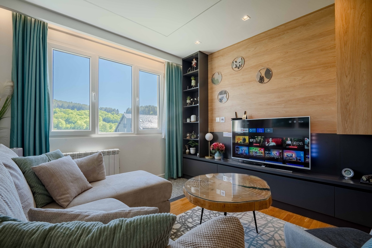 A stylish living area features large windows that provide ample natural light and offer views of the surrounding greenery. A comfortable sectional sofa sits beside a glass coffee table on a patterned rug, with a modern TV mounted on a wall adorned with decorative shelves.