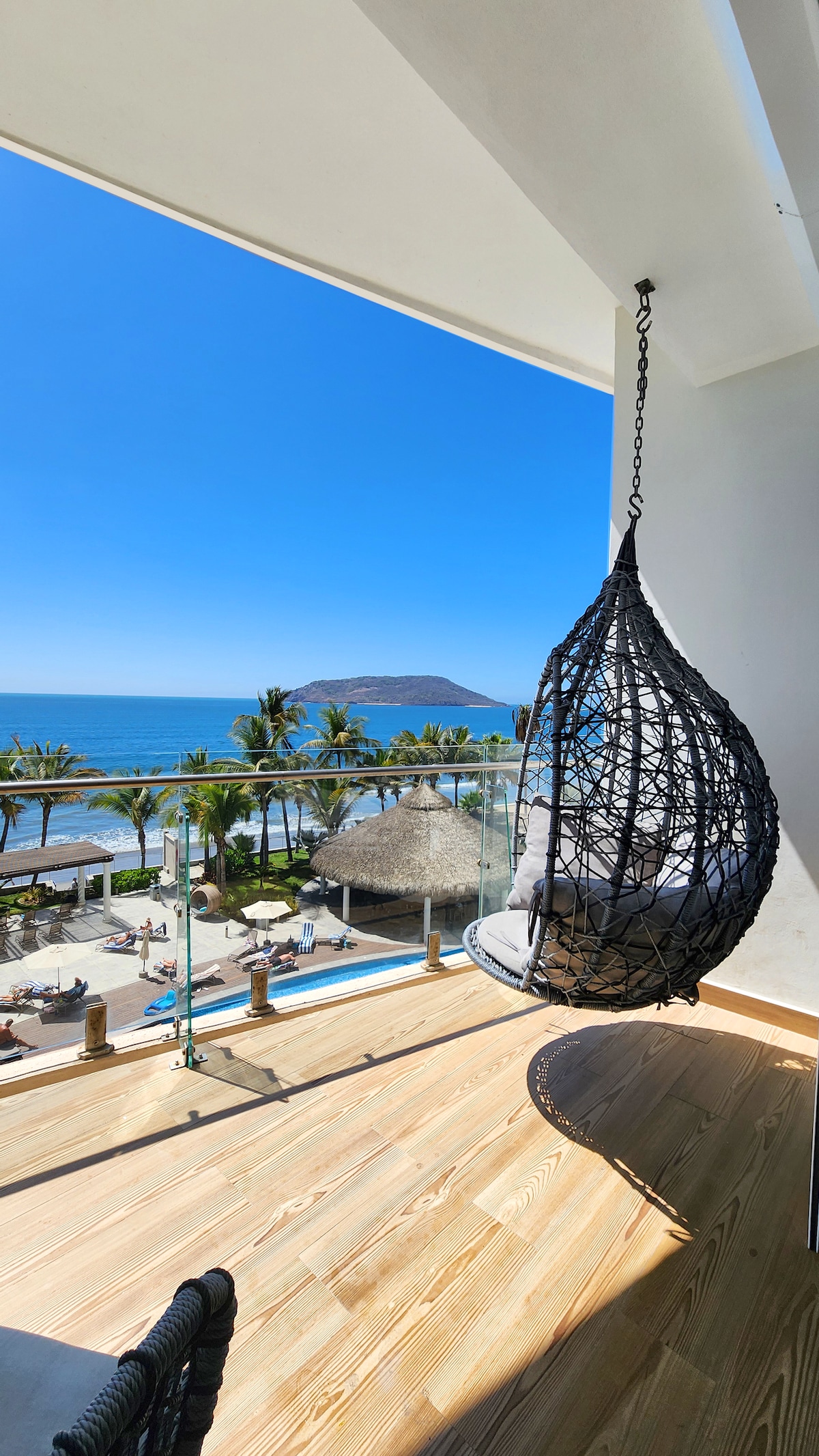 A suspended wicker chair is positioned on a balcony overlooking the ocean. Palm trees and a thatched-roof structure are visible below, while clear blue skies create a vibrant backdrop.