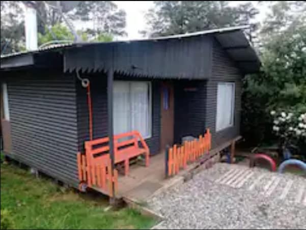 A small, rustic cabin with dark wooden siding is shown, featuring a sloped roof and a welcoming entry. A wooden bench sits on the porch, positioned beside well-maintained gravel pathways and surrounded by lush greenery.