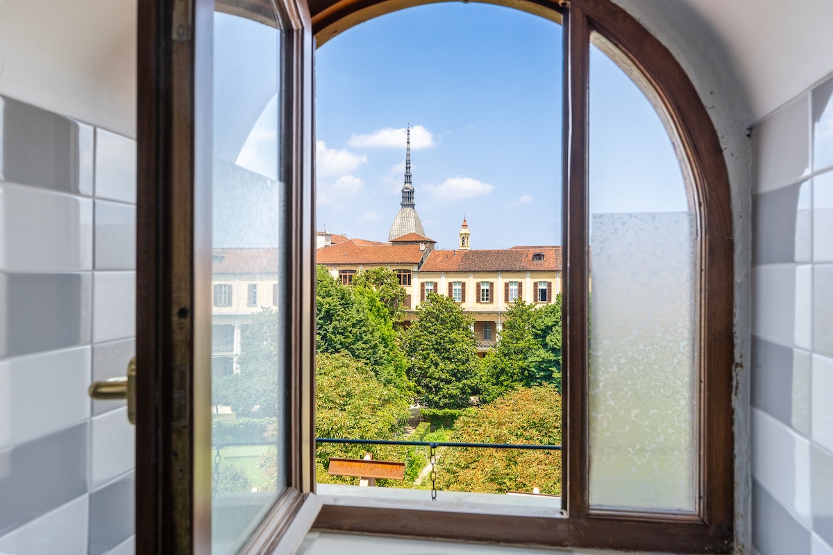 A window offers a view of lush greenery and historical buildings. The scene features a prominent tower in the background, framed by the arch of the window. The sunlight casts a soft glow on the outer garden area, enhancing the tranquil ambiance.