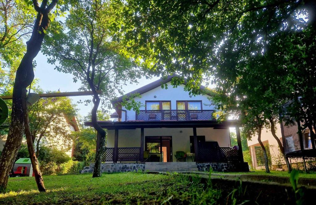 A two-story house is framed by lush green trees and a well-maintained lawn. The warm exterior is complemented by a large balcony with decorative railings. Sunlight filters through the foliage, creating a serene ambiance around the entryway.