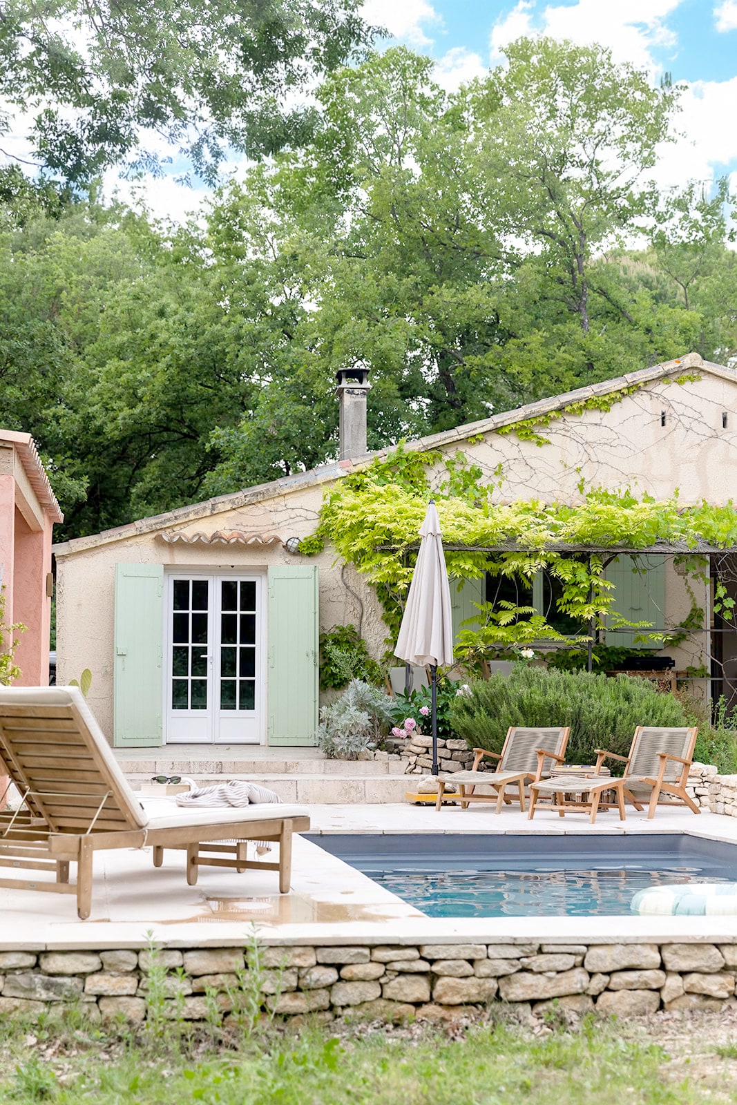 A charming villa is set amidst lush greenery, featuring a small pool surrounded by sun loungers. The façade showcases light green shutters and a traditional stone wall. A patio umbrella offers shade, enhancing the outdoor relaxation area by the villa.