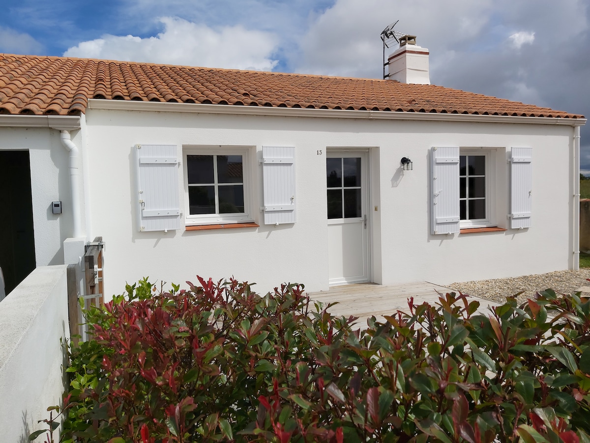 A single-story house with white exterior walls and terracotta roof tiles is shown. Windows with white shutters frame the entrance, which is equipped with a simple door. A green hedge with flowering foliage is visible in the foreground, enhancing the welcoming appearance of the property.