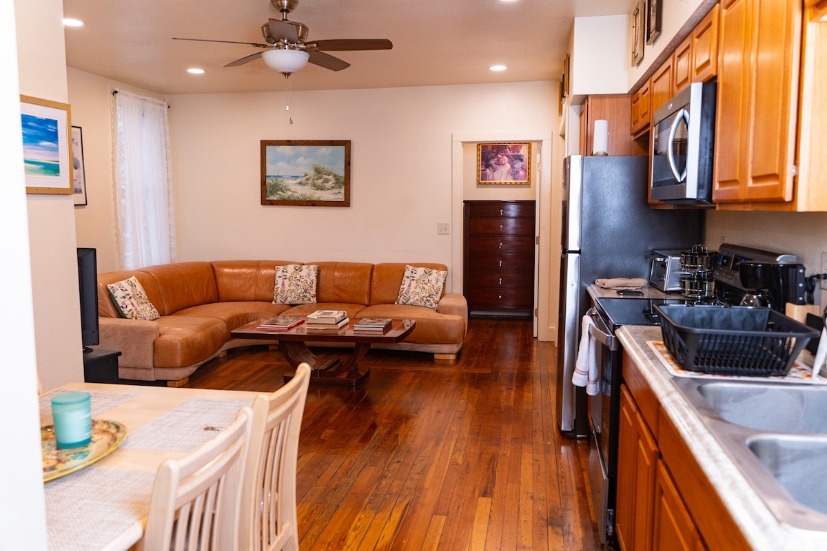 An open layout features a cozy living area with a curved leather sofa adorned with decorative pillows. The kitchen, equipped with modern appliances, is visible alongside a dining table set for four. Natural light brightens the space through nearby windows and decorative art pieces adorn the walls.