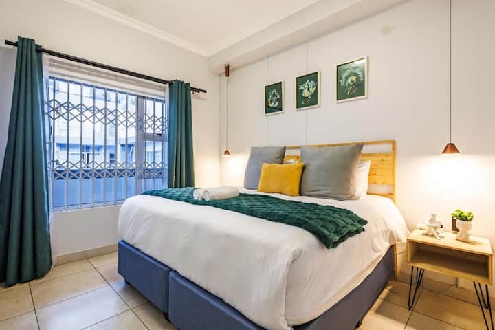 Charming and restful bedroom featuring a plush bed with crisp white linens, green and yellow accents, and curated artwork. Soft lighting and thoughtful décor create a serene space to relax and recharge.