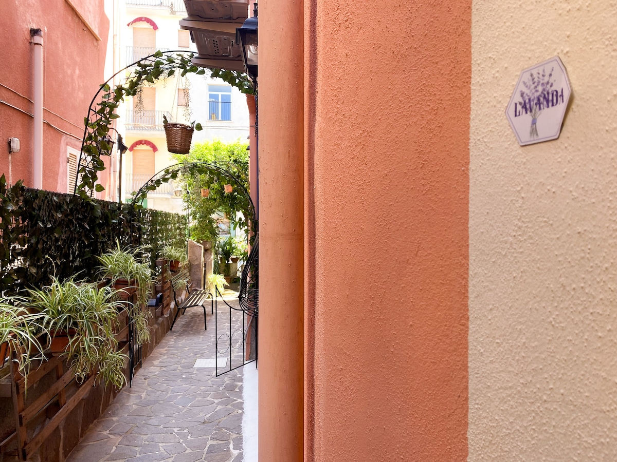 A quiet pathway is framed by vibrant greenery and terracotta-colored walls, leading to a small seating area. The pathway features arches adorned with hanging plants, inviting a sense of serenity in the surroundings.