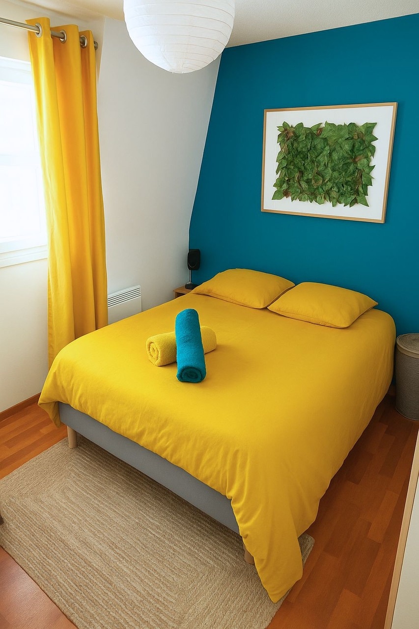 A cozy bedroom is adorned with a bright yellow bedspread and pillows. A blue accent wall contrasts with the warm tones, while a piece of wall art features leafy greenery. Towels are neatly arranged on the bed, and light filters through a nearby window, enhancing the inviting space.