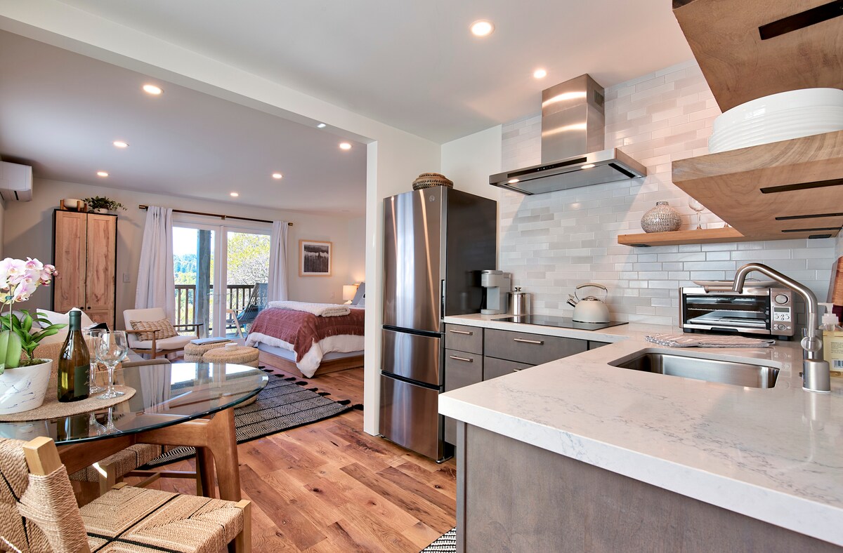 A modern, open-plan kitchen features sleek appliances and a marble countertop, complemented by wooden shelving. A cozy dining area with a glass table and woven chairs is visible, alongside a spacious living area with large windows, allowing natural light to fill the space.