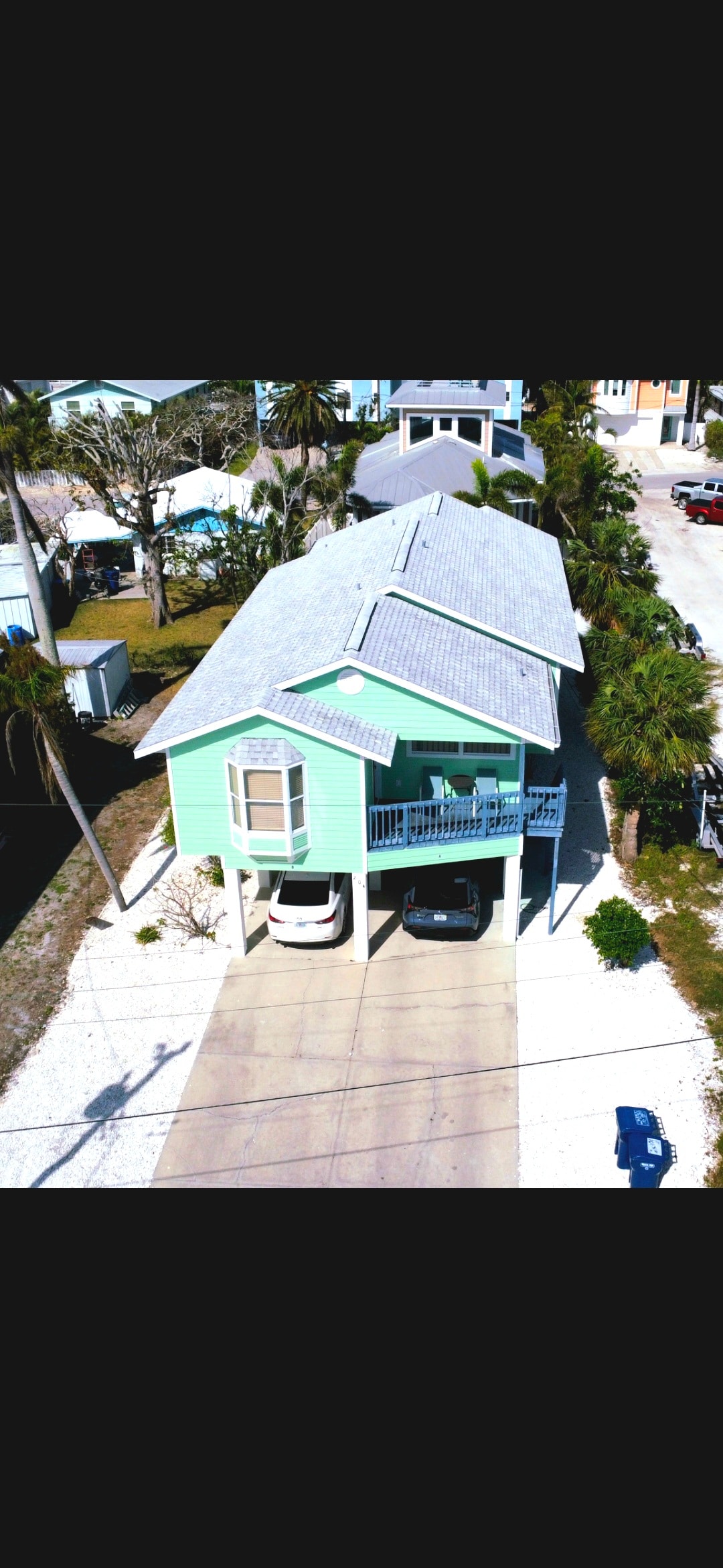 An aerial view of a newly renovated duplex showcases its light green exterior. The structure is positioned raised above ground, with a driveway accommodating multiple vehicles. Lush palm trees and a sandy path flank the property, providing a coastal atmosphere.