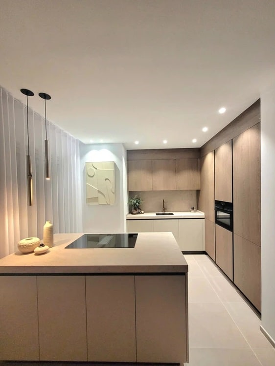 A modern kitchen features sleek cabinetry in warm wood tones, complemented by a minimalist island with an integrated cooktop. Soft lighting illuminates the space, highlighting the elegant design and neutral color palette, while a piece of wall art adds a subtle touch of sophistication.