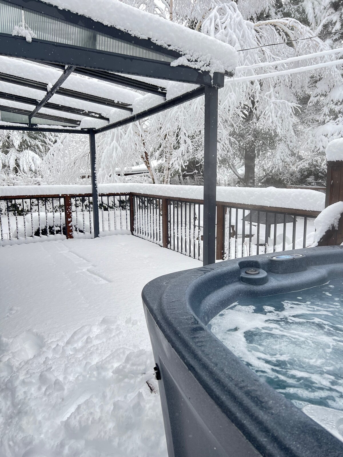 A hot tub is visible in the foreground, surrounded by a snow-covered deck. The roof overhead offers shelter from the elements. Fresh snow blankets the ground, creating a serene winter landscape. Trees in the background are also coated in snow, enhancing the tranquil setting.