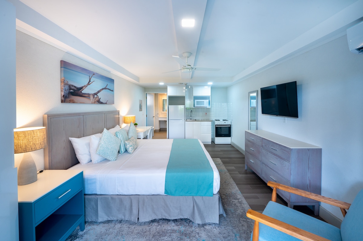 A spacious studio apartment is presented with a king-sized bed, featuring light bedding and decorative pillows. A kitchenette and living area are visible in the background, with modern furnishings, including a flat-screen TV and a comfortable wood chair. Soft lighting enhances the welcoming atmosphere.