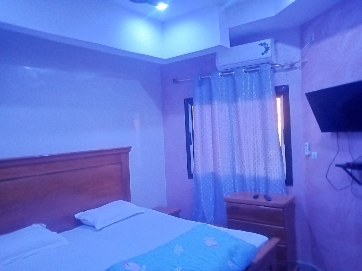 A serene bedroom features a spacious bed with soft linens, complemented by a light blue blanket. Natural light enters through a window covered with sheer curtains. A wooden dresser and a wall-mounted television enhance the room's functionality, framed by light purple walls.