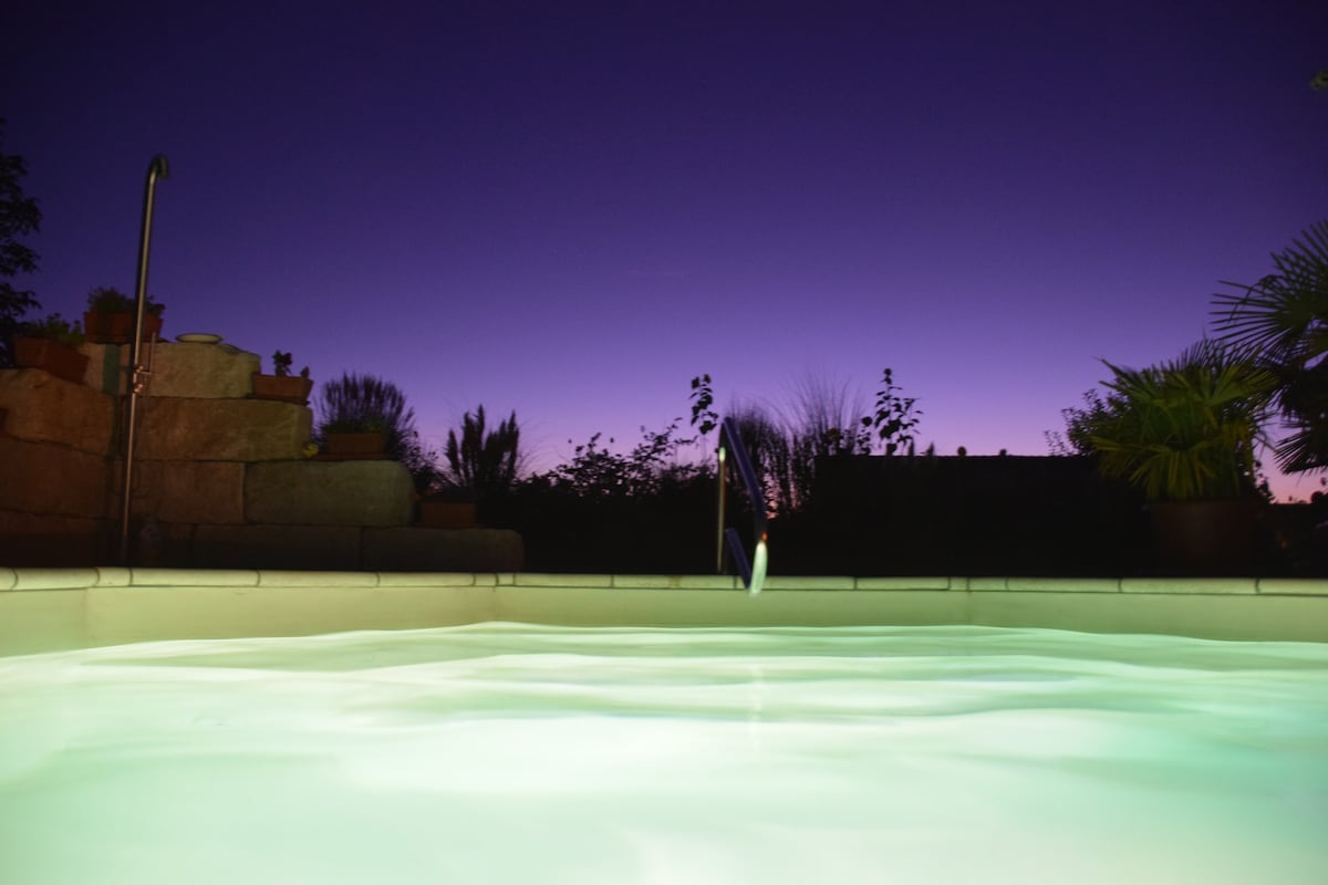 The image captures a serene outdoor swimming pool at dusk, illuminated with soft green lights. The calm water reflects the gradient of the evening sky, transitioning from deep blue to purple. Lush plants and stone steps create a tranquil atmosphere in the background.