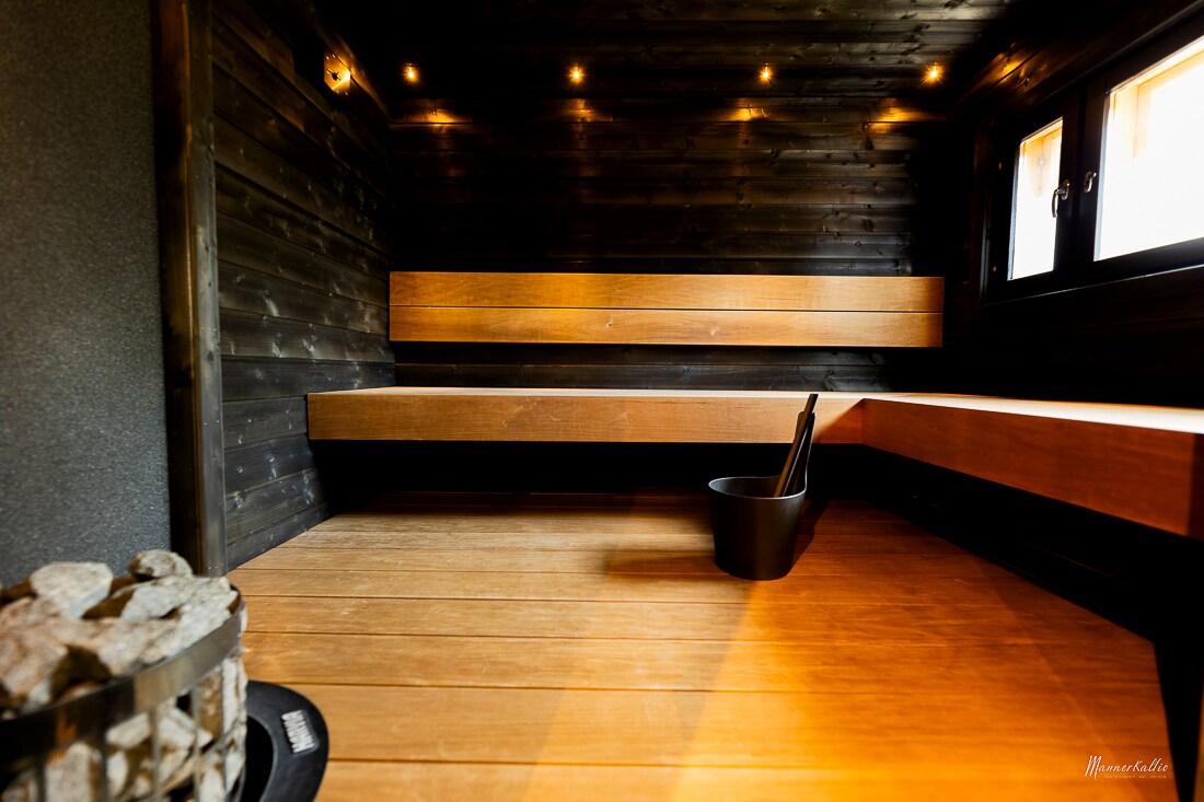 The interior of a spacious sauna is highlighted by wooden benches along the walls, complemented by subtle lighting. A traditional sauna heater is visible, with a bucket and ladle placed nearby. Large windows add natural light to the serene environment.