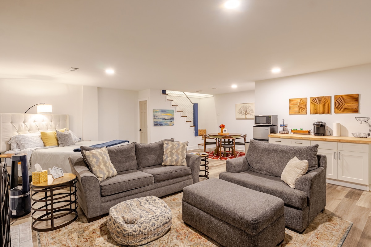 A modern suite is presented with a California king bed, gray sofa, and an oversized chair. A coffee table and kitchenette are positioned alongside, featuring a microwave and coffee maker. Soft lighting reflects off the neutral walls, creating a cozy, inviting setting.