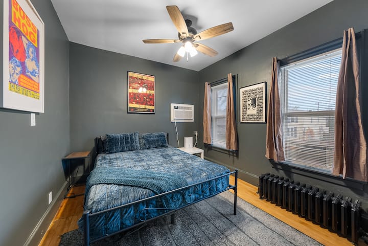 Deze kamer heeft een gezellige, rustgevende sfeer met diepgroene muren en zacht, gefilterd natuurlijk licht vanuit twee ramen. De warme gloed van de plafondventilator draagt bij aan het uitnodigende gevoel, waardoor een rustige en intieme ruimte ontstaat die ideaal is voor rust en ontspanning.