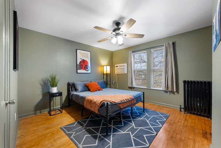 De groene slaapkamer voelt sereen en uitnodigend aan, met een rustgevende, natuurlijke sfeer. De hardhouten vloeren voegen warmte toe, terwijl het donkerblauwe tapijt voor een gezellig tintje zorgt. Het zwarte metalen bedframe en moderne lokale kunstwerken geven de kamer een strakke, artistieke energie.