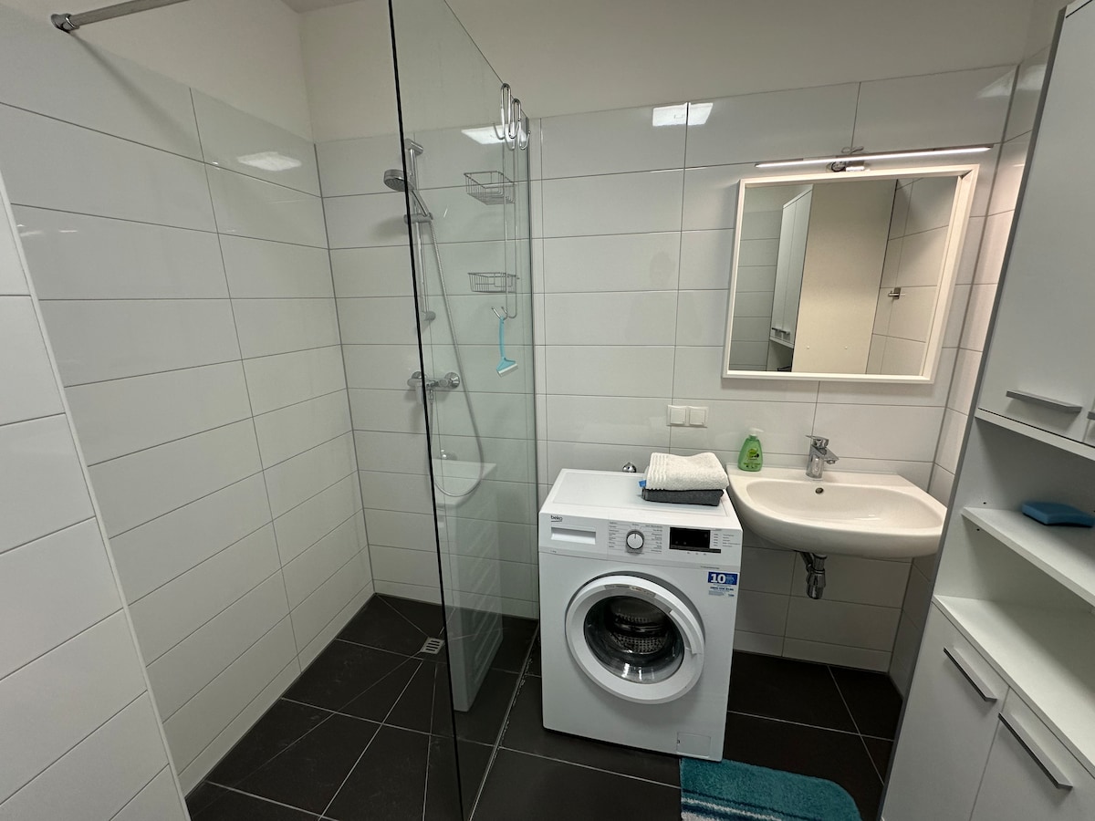 A modern bathroom features a glass shower enclosure and a washing machine. A mirrored vanity with a sink is positioned beside a storage unit. White tiles create a fresh and clean atmosphere, complemented by dark flooring. Soft lighting is provided by an overhead fixture.