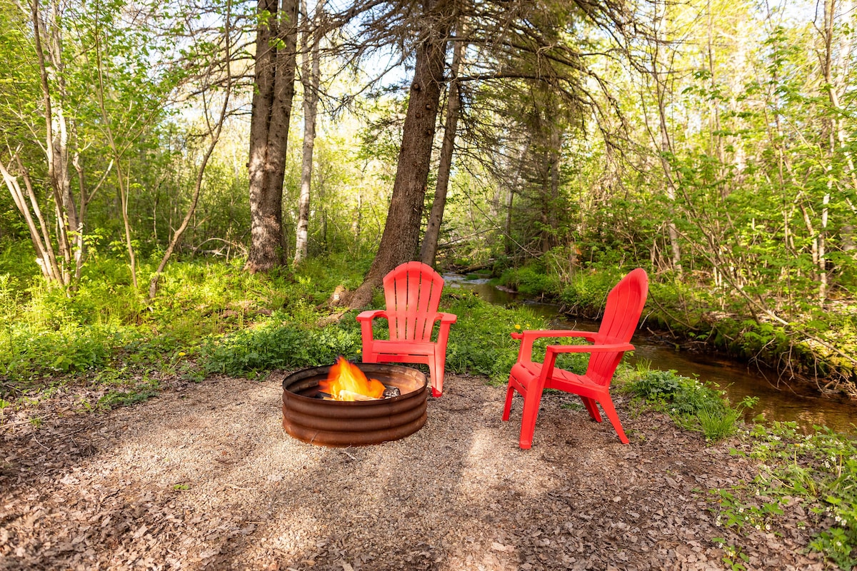 A serene outdoor fire pit area is surrounded by lush greenery and trees. Two red Adirondack chairs are positioned near a circular stone fire pit, which is lit with a warm fire. A tranquil stream runs alongside the seating area, enhancing the natural setting.