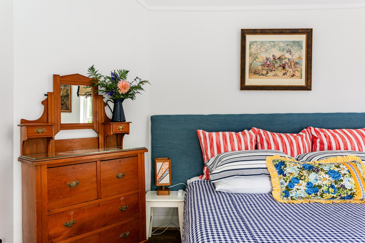 A cozy bedroom area features a spacious bed adorned with striped and floral bed linens. A vintage wooden dresser is positioned nearby, accented by a floral arrangement and framed artwork hanging above the bed. Soft light illuminates the room, enhancing its serene ambiance.