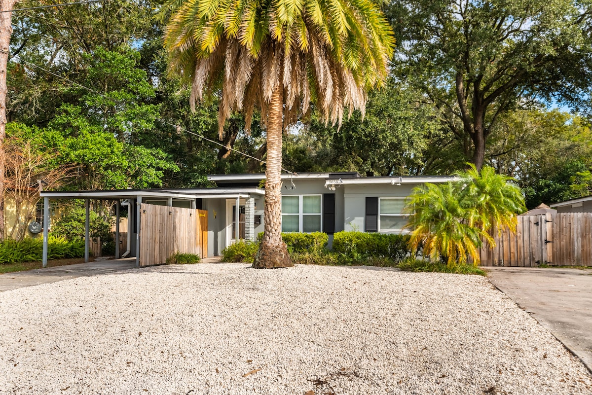 A single-story guest suite is framed by lush greenery and a large palm tree. The entrance is highlighted by a covered carport and a gravel driveway, providing a welcoming approach. Large windows invite natural light, complementing the tranquil surrounding landscape.