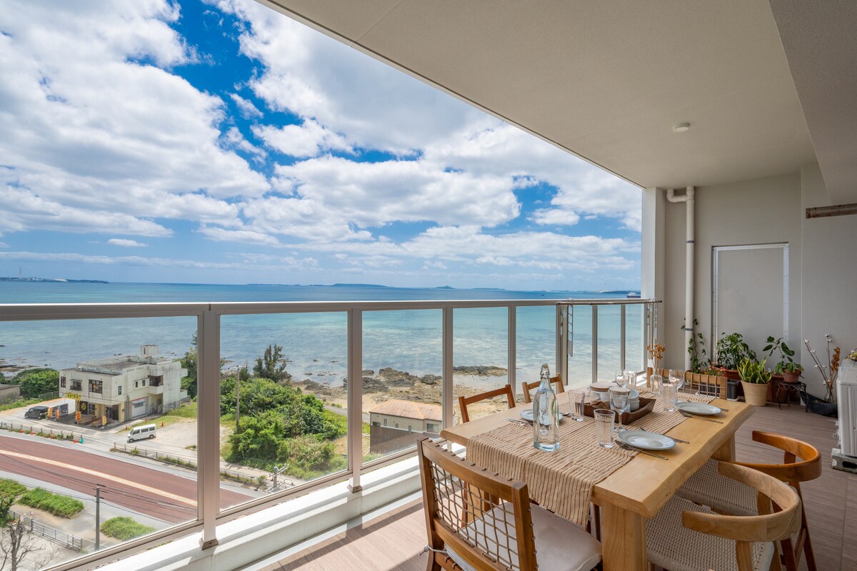 A spacious balcony offers a dining table set for four, with an ocean view in the background. The scene is complemented by light clouds under a blue sky, presenting a serene coastal atmosphere. A road and lush greenery can be seen below.