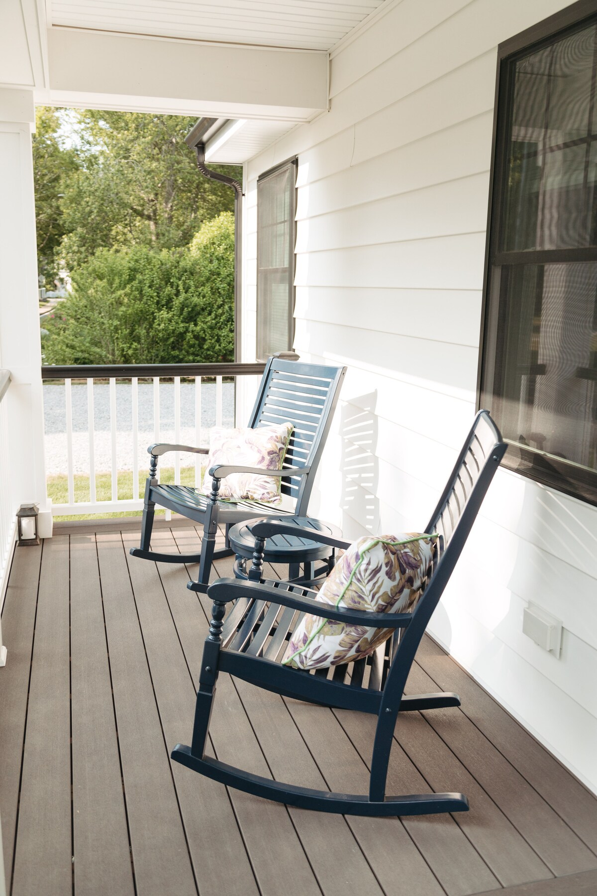 A front porch area is shown with two blue rocking chairs, each adorned with patterned cushions. The flooring is wooden, and sunlight filters through large windows, creating a serene outdoor space for relaxation.