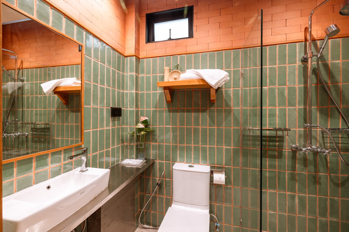 A modern bathroom is presented with green tiled walls, complemented by a sleek vanity. Natural light is filtered through a small window. A glass shower enclosure is visible alongside a clean toilet and well-organized shelving that holds towels and decorative items.