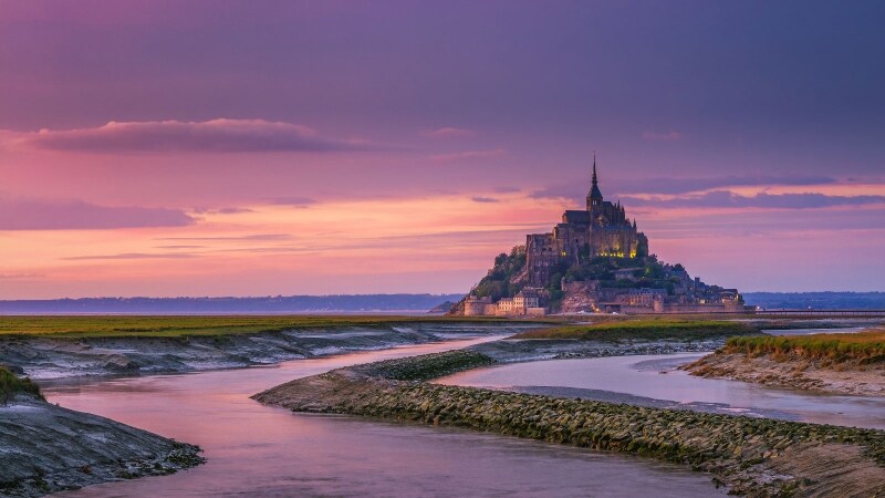A serene view features a historic island structure, set against a twilight sky with soft shades of purple and orange. A winding river flows through the grassy foreground, reflecting the natural beauty of the surroundings and leading towards the iconic silhouette of the island.