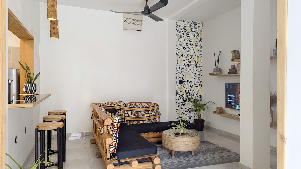 A bright living area features a unique wooden seating arrangement with colorful cushions. A natural fiber coffee table sits on a woven rug, while a wall-mounted television is visible. Green plants are placed throughout the space, and a ceiling fan is positioned above.