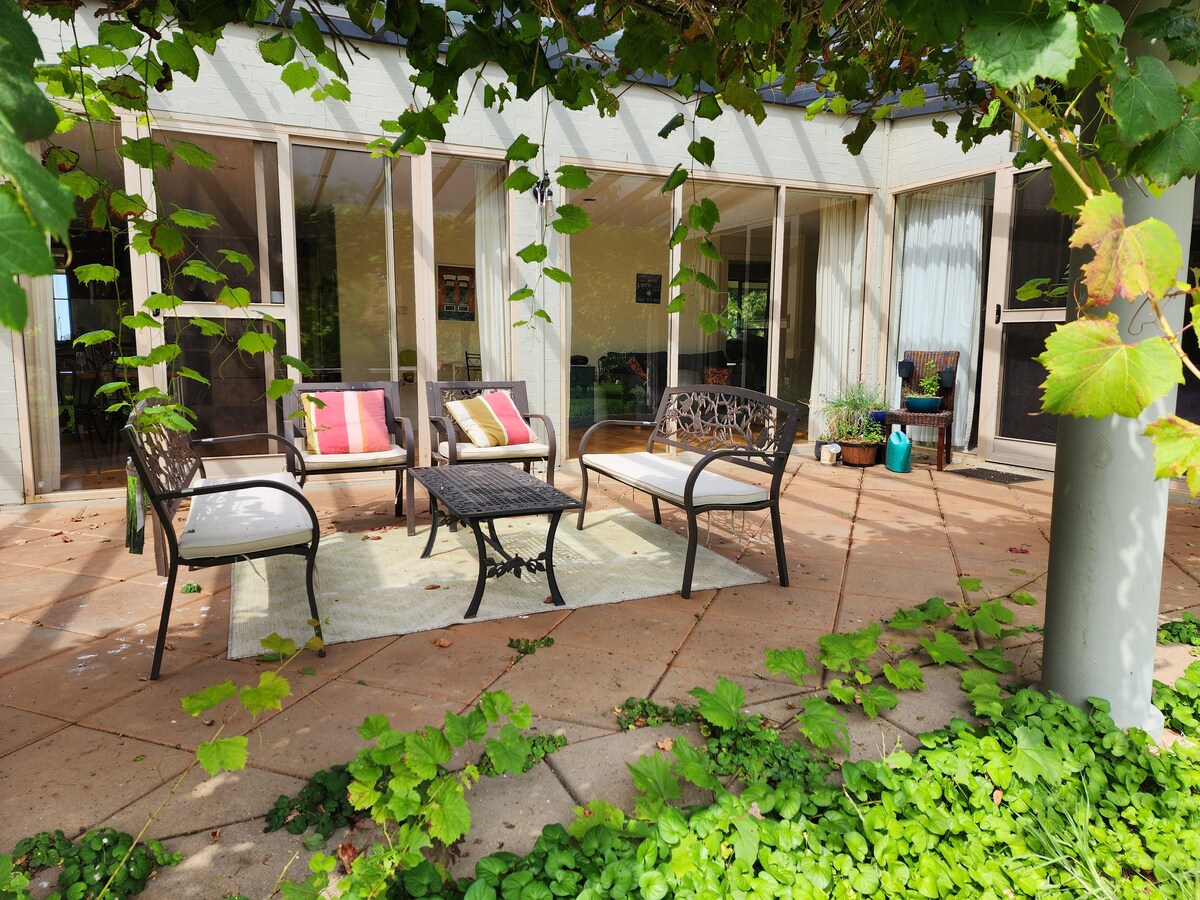 A shaded outdoor seating area features two cushioned chairs and a sofa arranged around a central table. The space is adorned with climbing vines and potted plants, with natural light illuminating the comfortable setting.