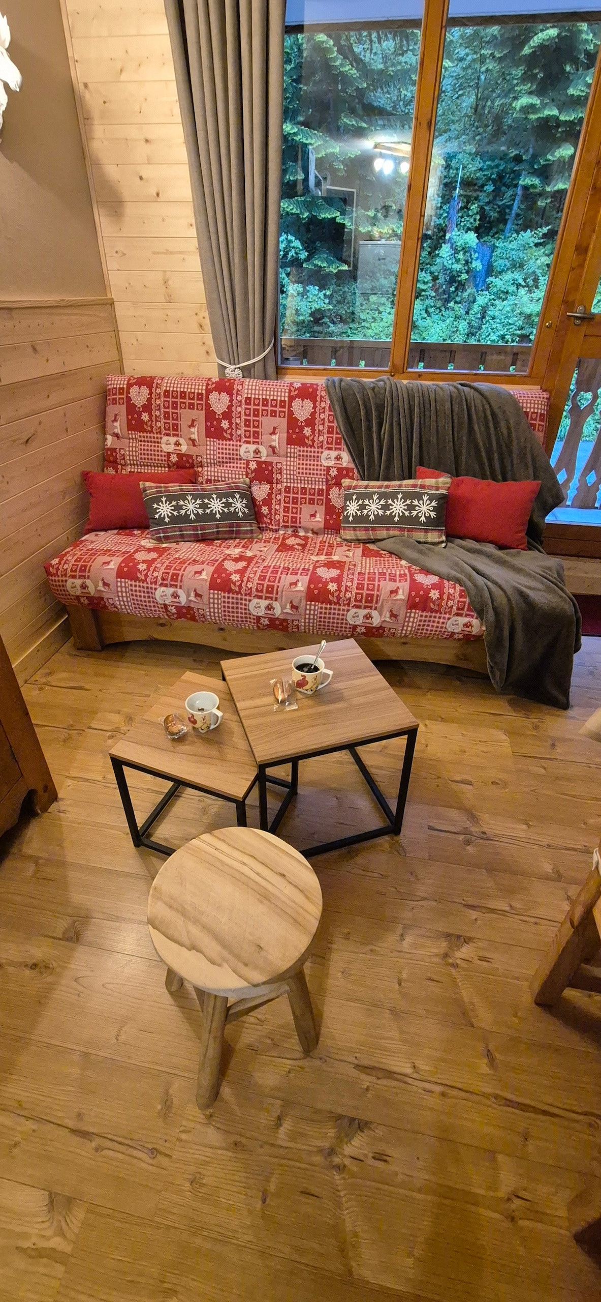 A comfortable seating area is equipped with a red and white patterned sofa adorned with decorative pillows. Two wooden coffee tables are positioned in front, accompanied by a small stool. Large windows provide a view of the greenery outside, illuminated by natural light.
