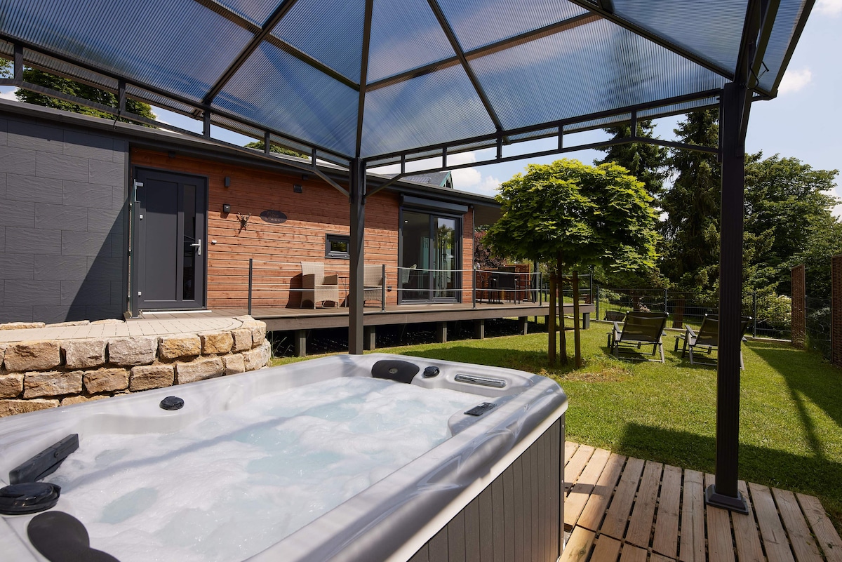 A covered outdoor hot tub sits on a wooden deck, surrounded by a grassy area and trees. The structure features semi-transparent roofing, providing shade while maintaining an open feel. In the background, a spacious terrace with seating is visible, enhancing the serene outdoor setting.