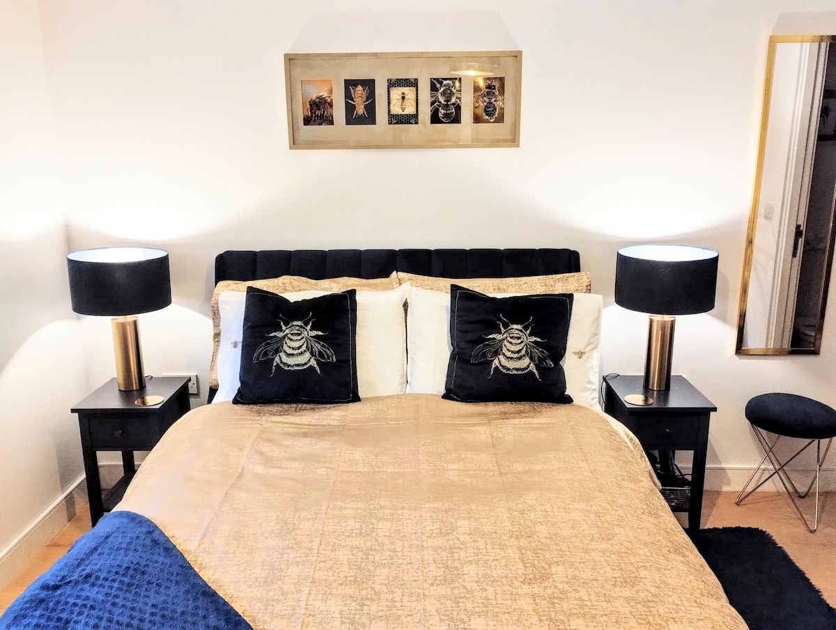 A double bed is adorned with rich gold and blue linens, flanked by two bedside tables with stylish lamps. Decorative bee-themed cushions add a unique touch. A framed art piece hangs above the bed, complemented by a nearby mirror reflecting the room's light.