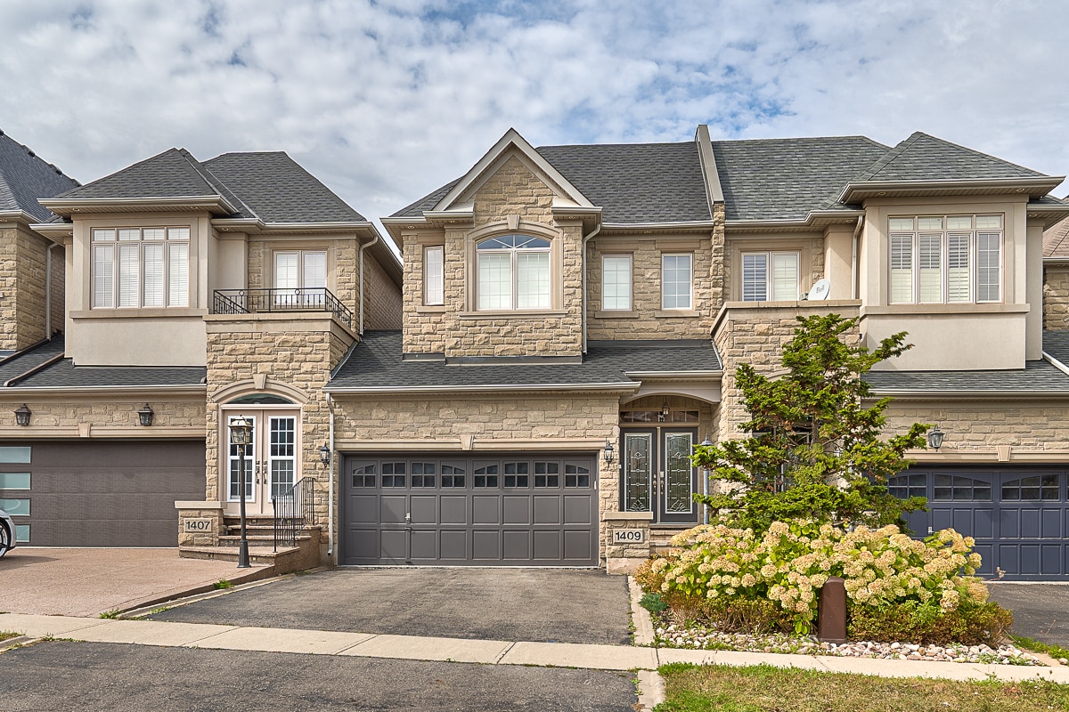 The exterior of a modern stone townhome features a double-car garage and an inviting entrance. Lush landscaping includes shrubs and ornamental plants, contributing to a well-maintained appearance. A cloudy sky hovers above, framing the stylish architecture of the neighboring homes.