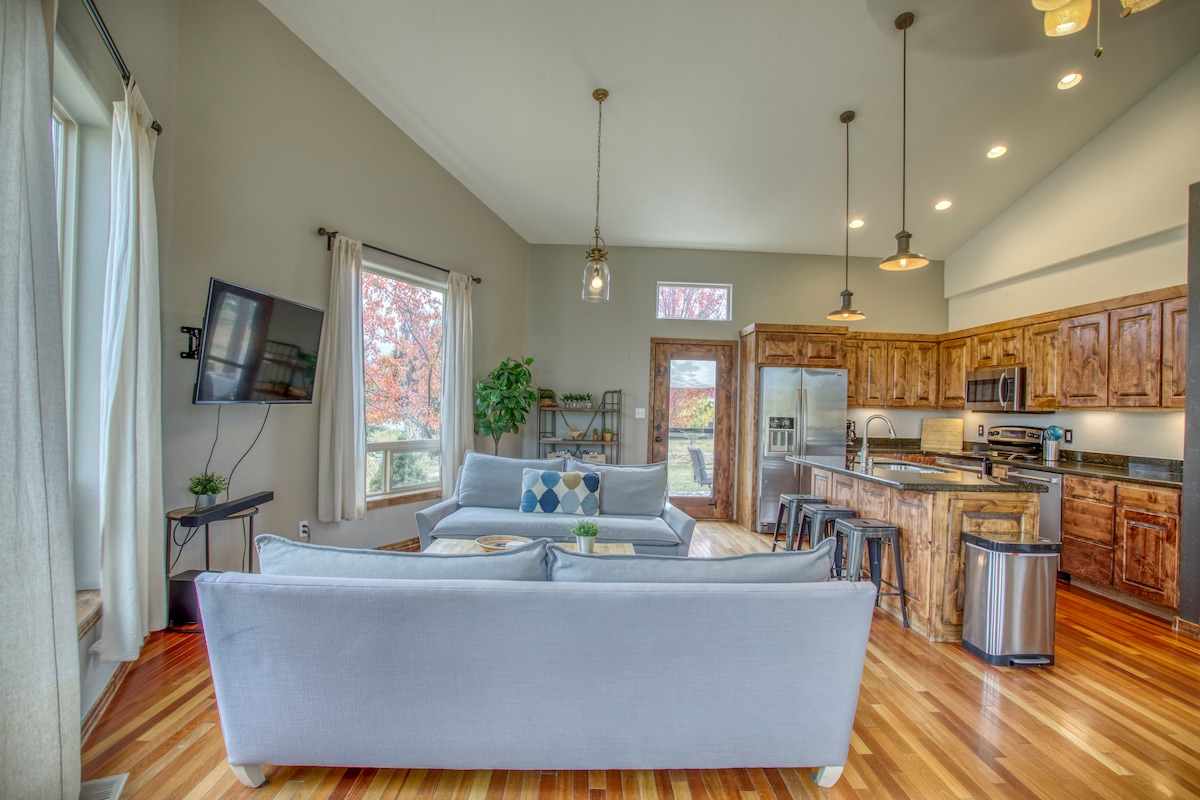 A spacious living area features large windows allowing natural light to fill the room. Comfortable seating is arranged near a wall-mounted television. The open layout connects to a kitchen with wooden cabinetry, stainless steel appliances, and an island with bar seating, enhancing the communal atmosphere.