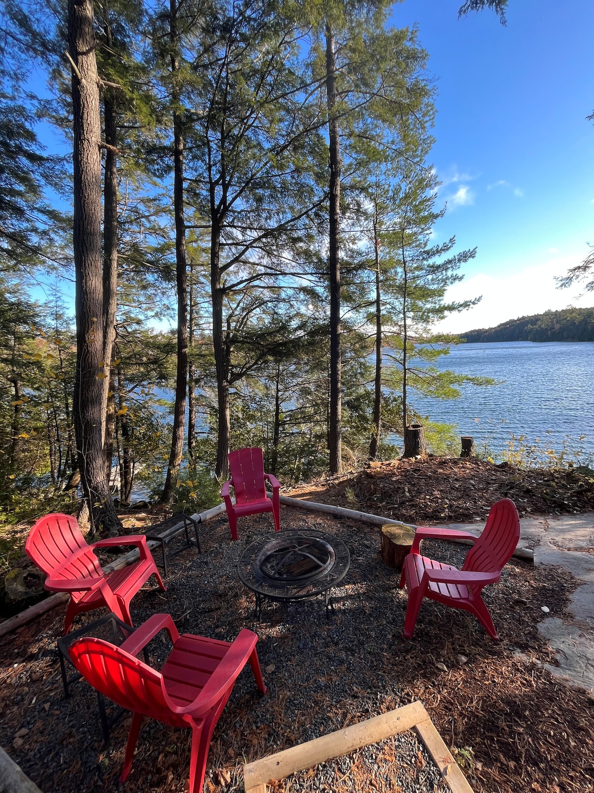 A relaxing outdoor fire pit area is surrounded by tall trees and designed for gatherings. Four red chairs are positioned around a circular stone fire pit. The serene lake is visible in the background, reflecting the clear blue sky and the gentle colors of nature.