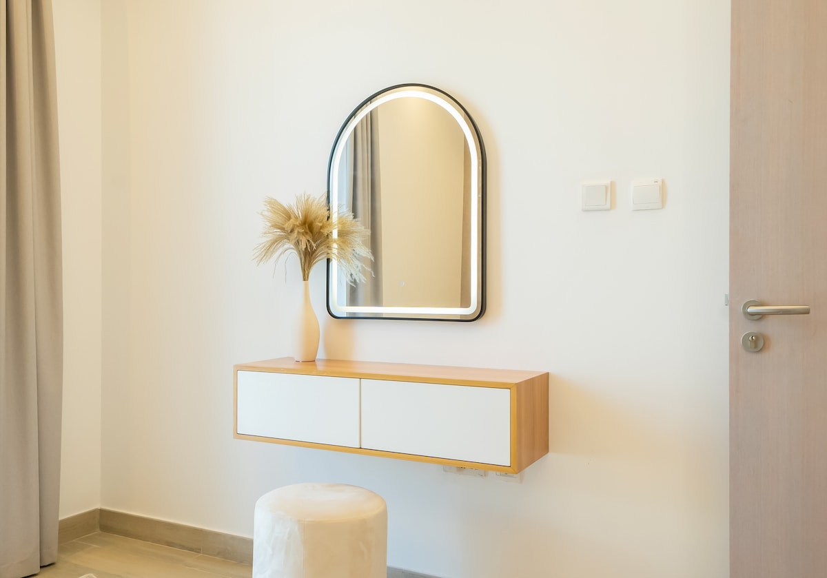 A minimalist entryway features a wall-mounted console with two drawers. A round, upholstered stool sits beneath it. Above, an arched mirror is framed in black, reflecting natural light, while a simple vase holds decorative dried grasses, adding a touch of warmth to the space.