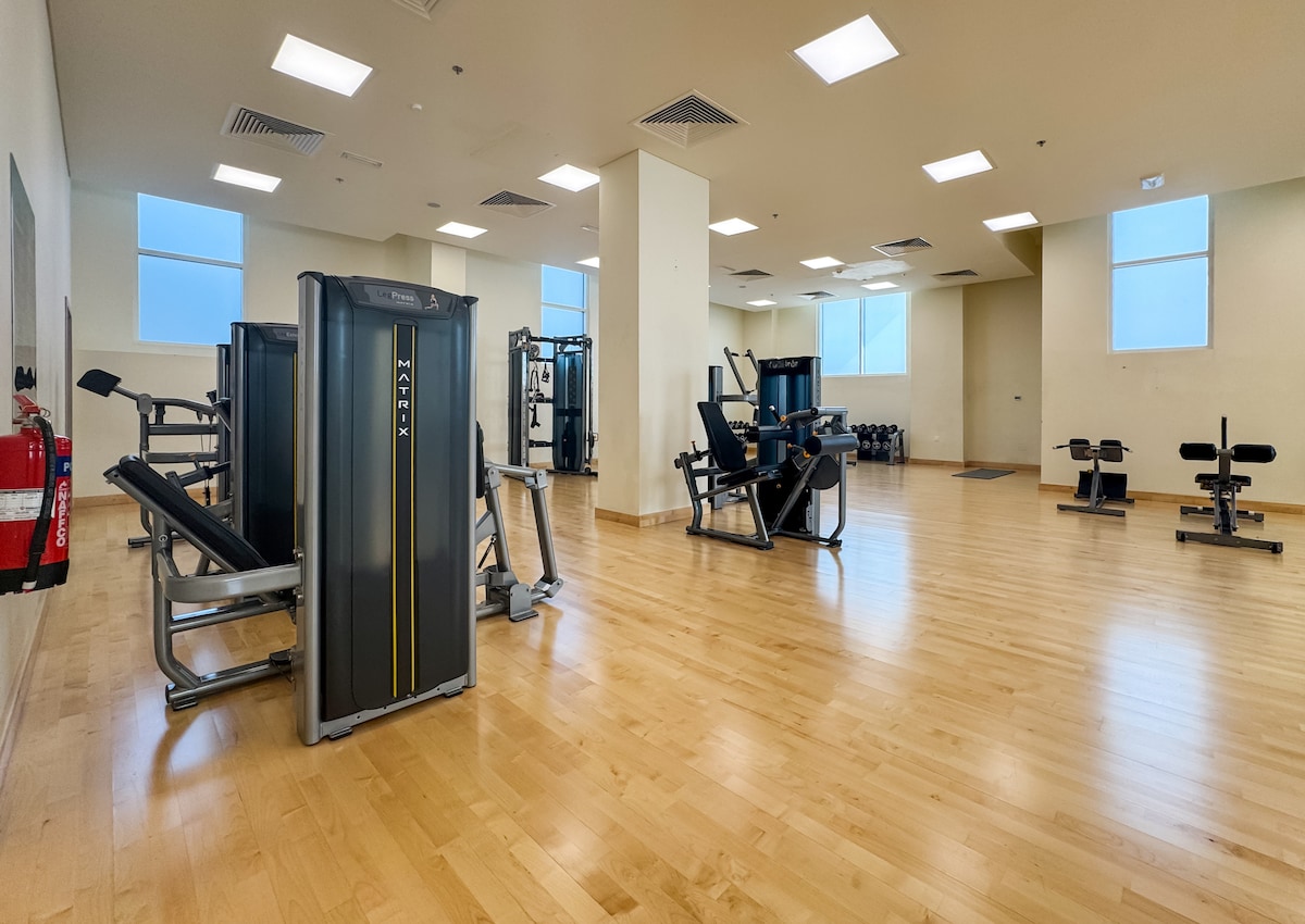 A modern gym space is showcased, featuring various fitness equipment arranged throughout a spacious room with wooden flooring. Large windows provide ample natural light, contributing to an open and inviting atmosphere. Wall-mounted mirrors enhance the sense of space and functionality.