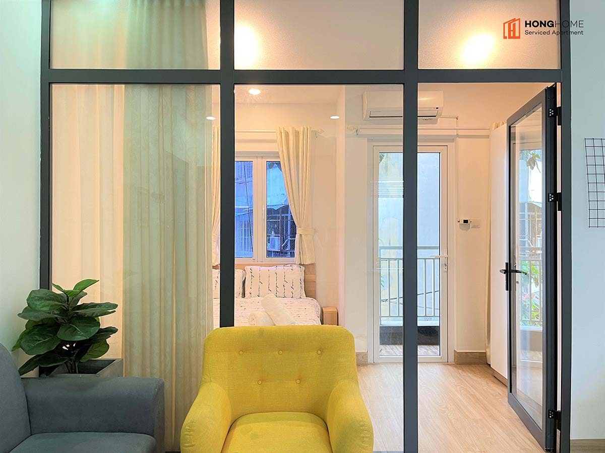 The living area is separated by glass sliding doors, allowing light to flow into the space. A gray sofa and a bright yellow chair are positioned in the foreground. Beyond the doors, a cozy sleeping area is visible, along with a balcony view that provides access to fresh air.
